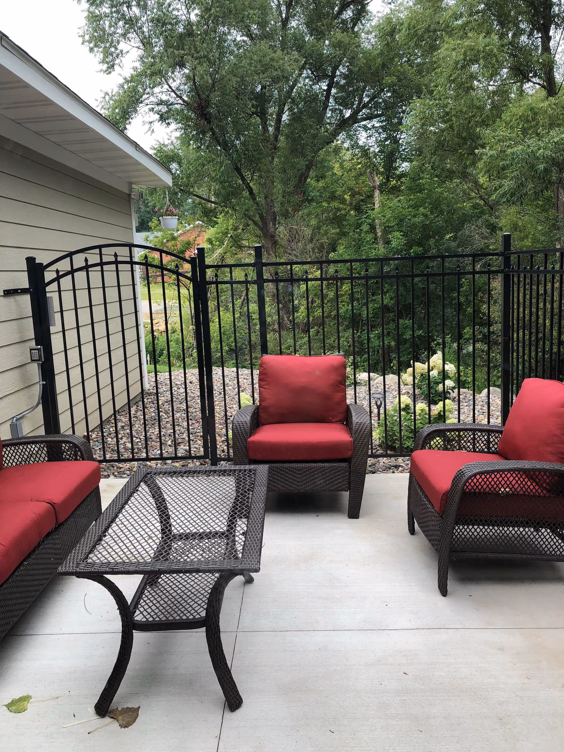 A patio with wicker seating and red cushions, a metal mesh coffee table, and a black fence overlooking trees.