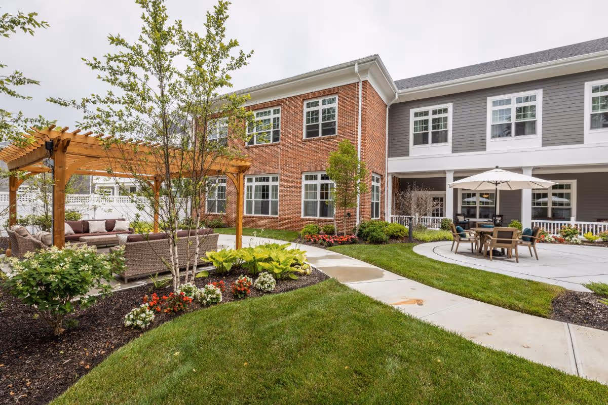 Outdoor courtyard at Sunrise of Huntington featuring a pergola with lounge seating, a dining table with umbrella, landscaped flower beds and a two-story brick-and-gray building.