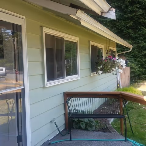 Exterior side view of a light green building with two windows and a sliding glass door. A black metal bench is placed on a small concrete patio area next to a wooden railing. Hanging flower baskets with pink and white flowers are attached under the roof eaves. Green grass and trees are visible in the background.