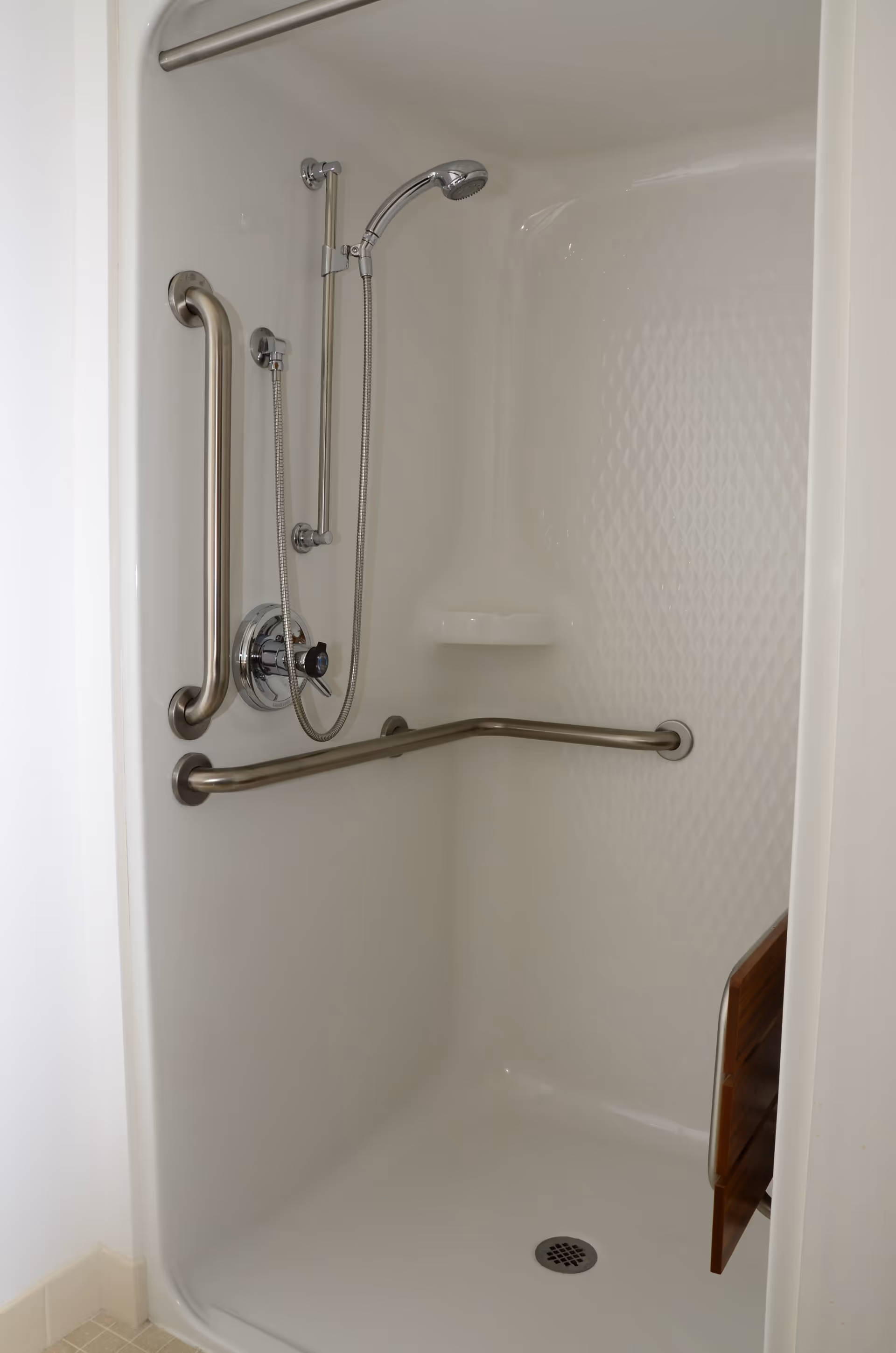 A white shower stall equipped with a handheld showerhead, two horizontal and one vertical stainless steel grab bars for support, and a foldable wooden shower seat attached to the wall.