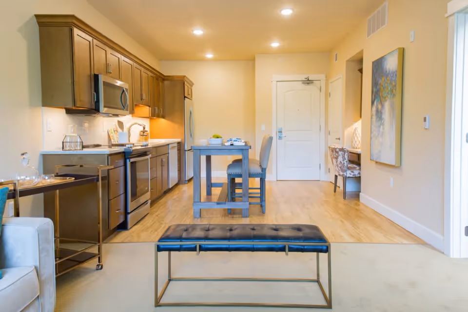 Open-plan apartment interior with a modern kitchen on the left, a small dining table in the center, and a bench in the foreground.