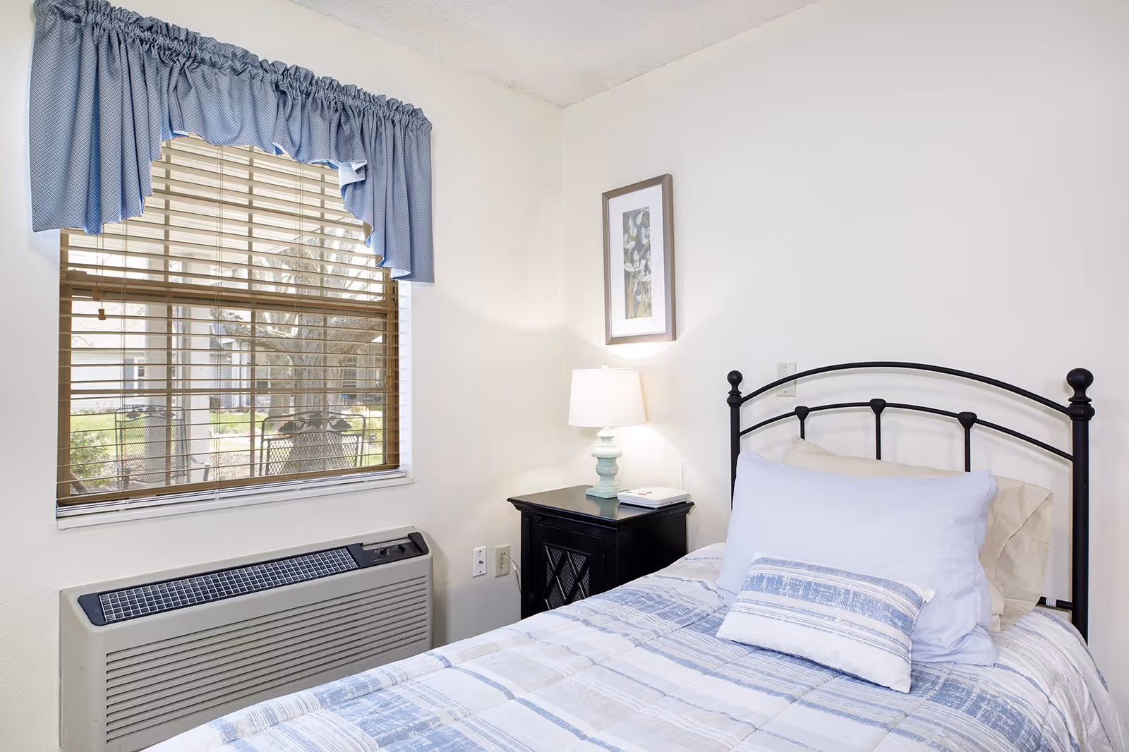 A small bedroom with a single bed featuring a black metal headboard, white and blue bedding, and two pillows. Next to the bed is a black nightstand with a light blue lamp and a book. A framed picture hangs on the white wall above the nightstand. A window with wooden blinds and a blue valance is on the left side, with an air conditioning unit below it.