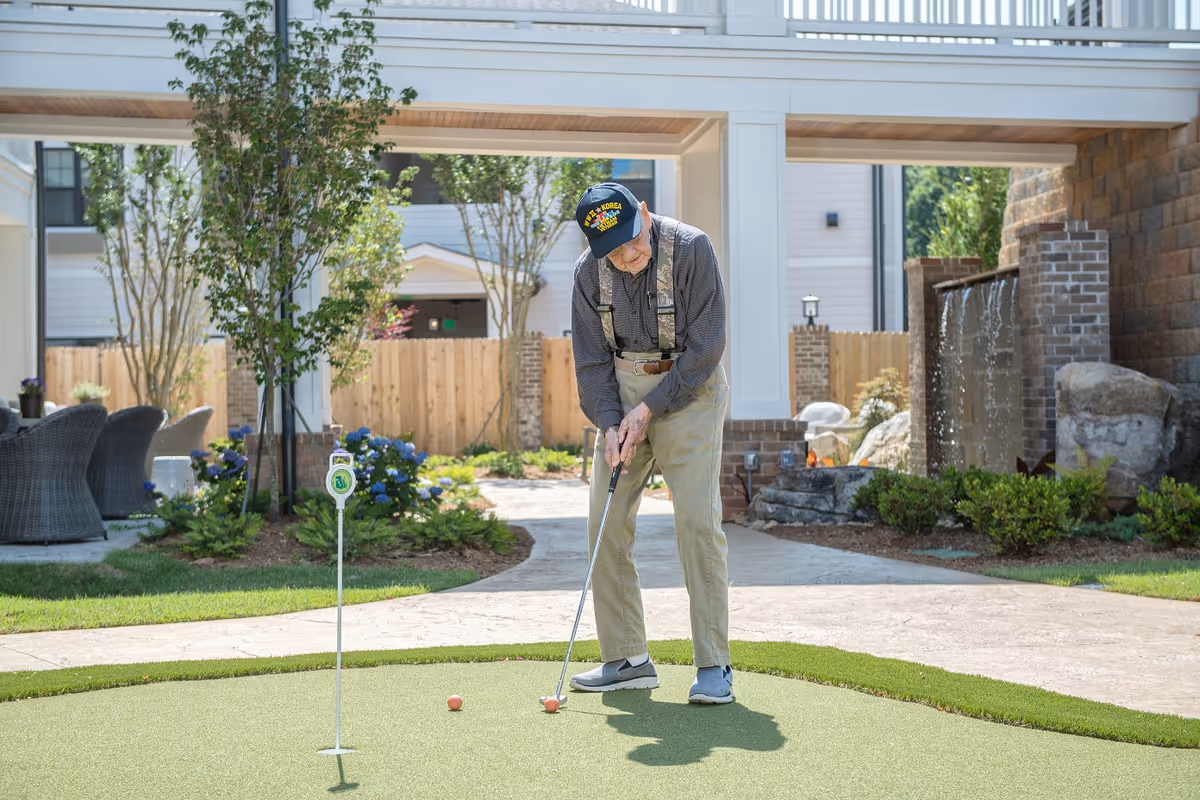 An elderly man wearing a Korea veteran cap and suspenders is playing putting golf on a small artificial green in an outdoor garden area with trees, plants, and a water feature in the background.