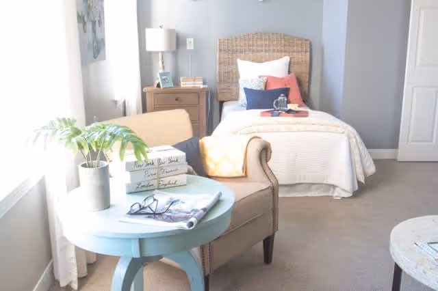 A bright and cozy bedroom with a single bed featuring a wicker headboard, white bedding, and colorful pillows. Next to the bed is a wooden nightstand with a lamp, books, and a framed photo. In the foreground, there is a light blue round table with a potted plant, stacked books, and a pair of glasses resting on an open magazine. A beige armchair is positioned beside the table, and sunlight streams in through a window with white curtains.