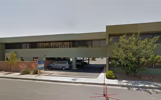 Exterior view of a two-story building with a covered parking area underneath. There are a few small trees and shrubs along the sidewalk in front of the building. A sign is placed near the curb on the left side of the image.