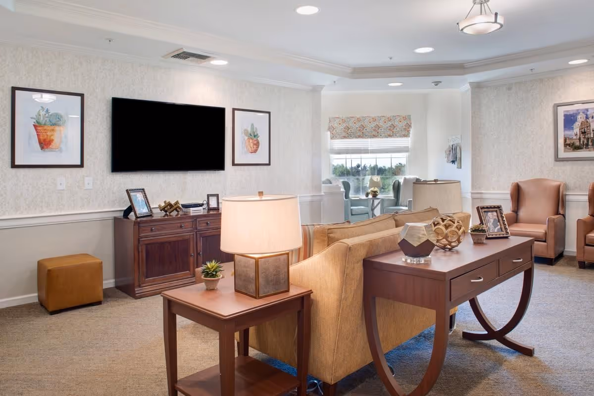 A cozy living room area in a senior living facility featuring a beige sofa, two brown armchairs, a wooden console table with decorative items and lamps, a wooden cabinet with framed photos, a wall-mounted flat-screen TV, and framed artwork on the walls. A window with a patterned valance lets in natural light, and the room has a neutral color palette with carpeted flooring.