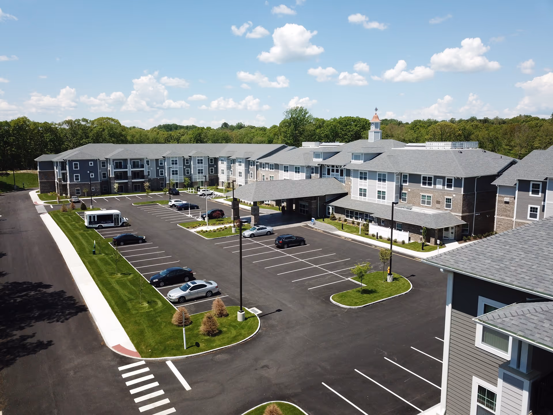Aerial view of a three-story retirement community building with a covered entrance, parking lot, and landscaped grounds under a blue sky.