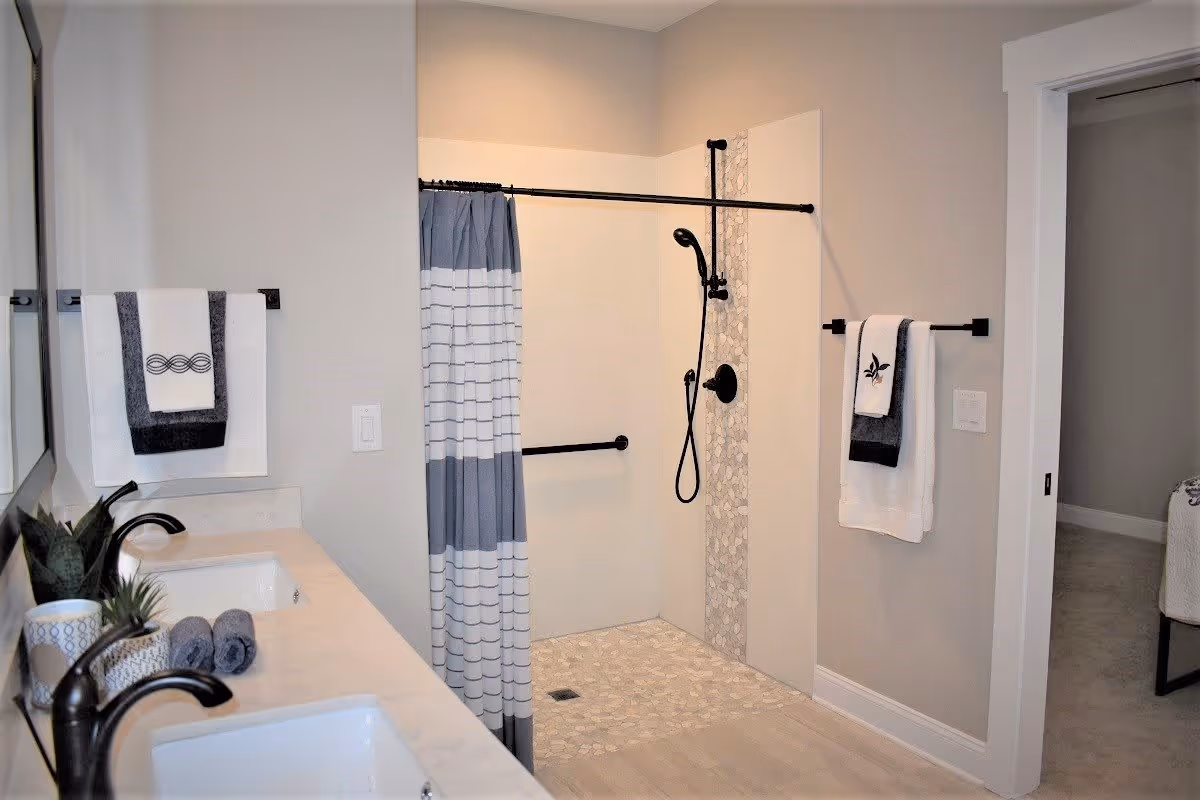 A modern bathroom featuring a double sink vanity with black faucets, a potted plant, and rolled towels on the countertop. The shower area has a black showerhead and grab bar, a patterned tile strip, and a blue and white striped shower curtain. Two towel racks hold neatly folded towels. The bathroom walls are painted light beige, and a doorway leads to an adjacent room.