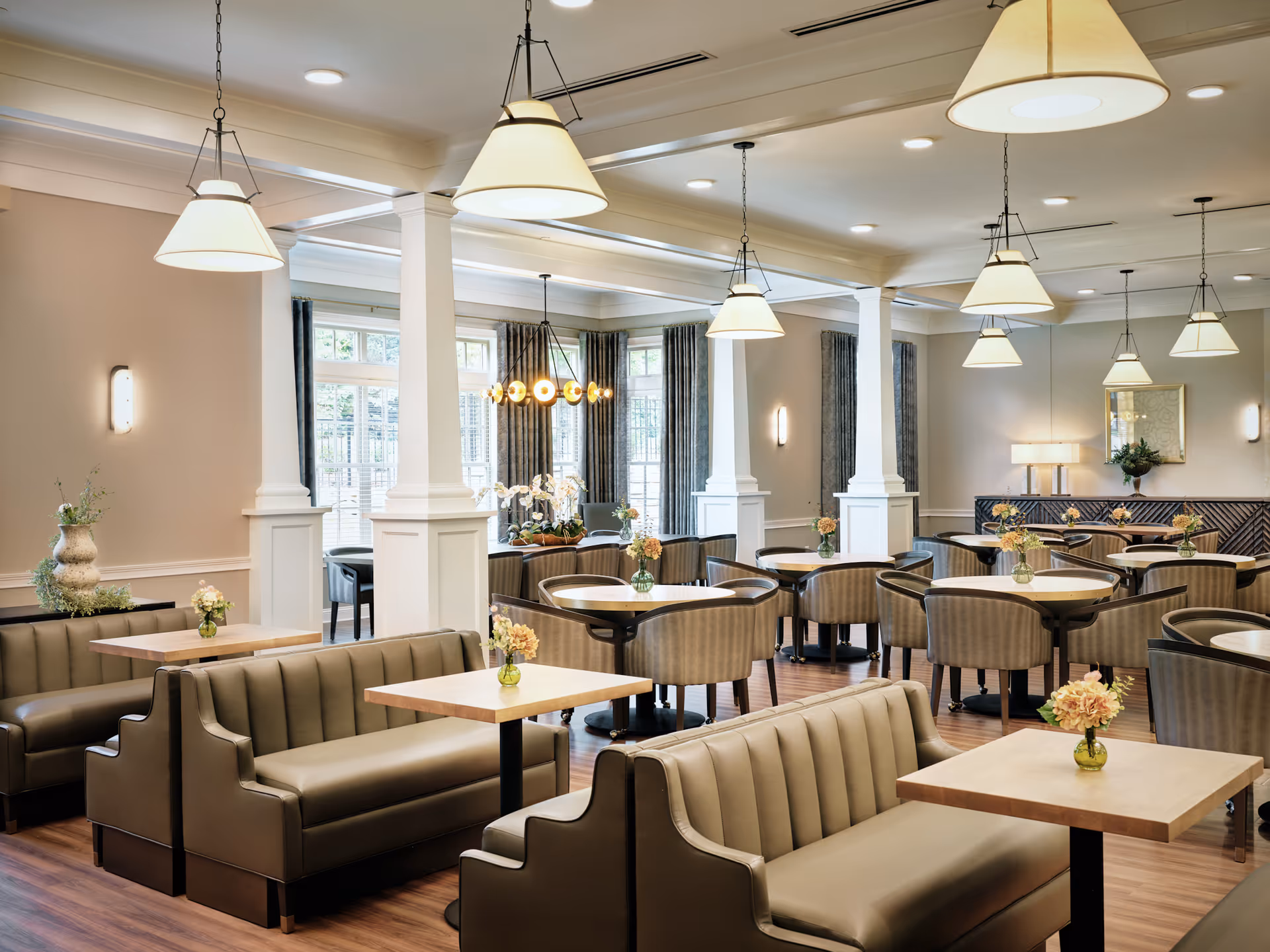 A spacious senior living dining room with multiple tables and chairs arranged neatly. The room features beige cushioned booth seating, round tables with striped upholstered chairs, and wooden floors. Large windows with gray curtains allow natural light to fill the space. Several pendant lights hang from the ceiling, and small vases with flowers decorate each table.