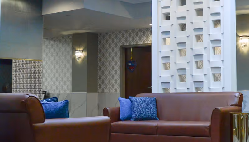 A cozy interior lounge area with brown leather sofas adorned with blue cushions. The background features patterned wallpaper, a decorative white partition, and a door with a hanging heart-shaped ornament.