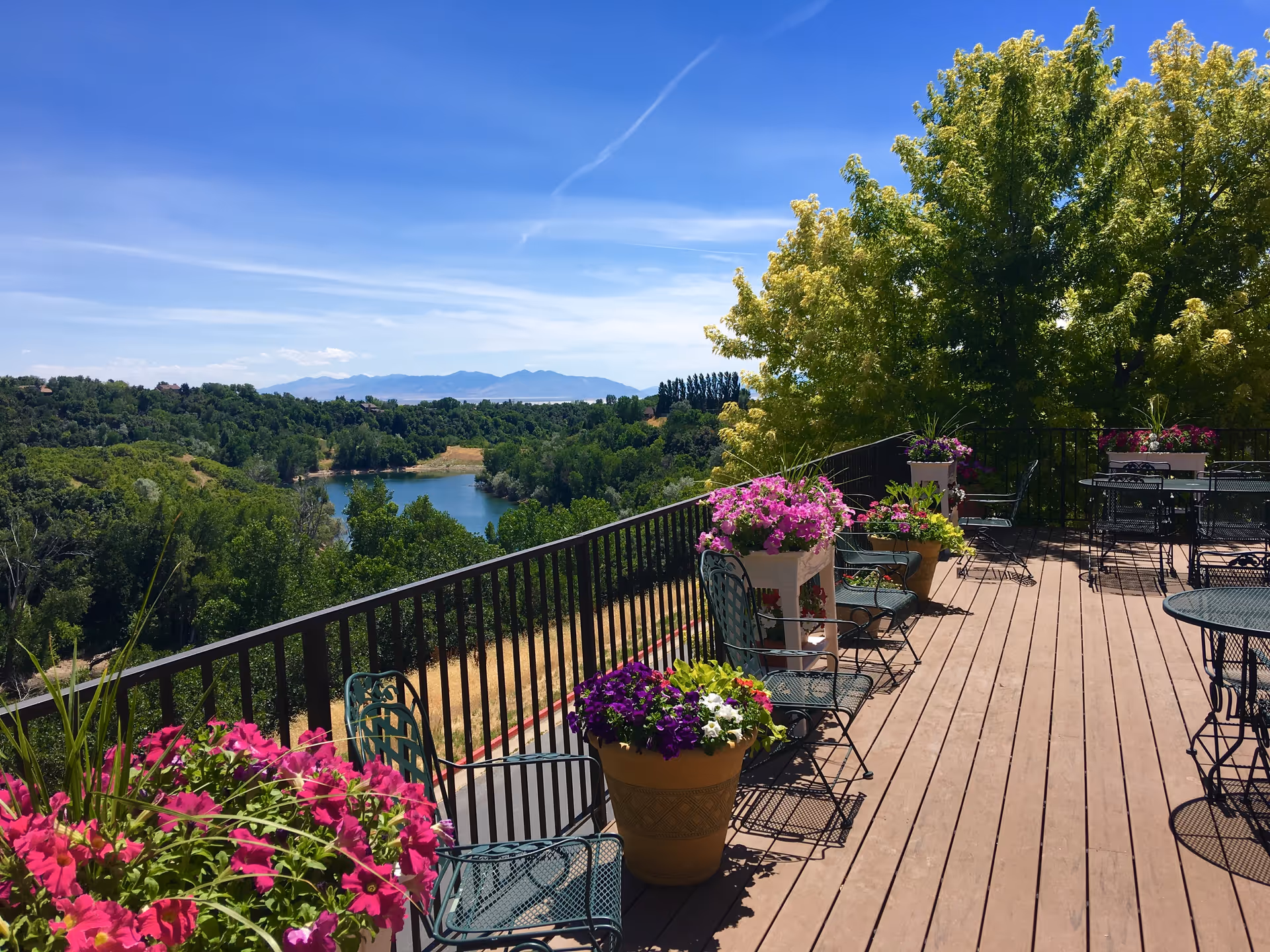 Sunlit outdoor deck with potted flowers, metal tables and chairs overlooking a lake and wooded landscape.