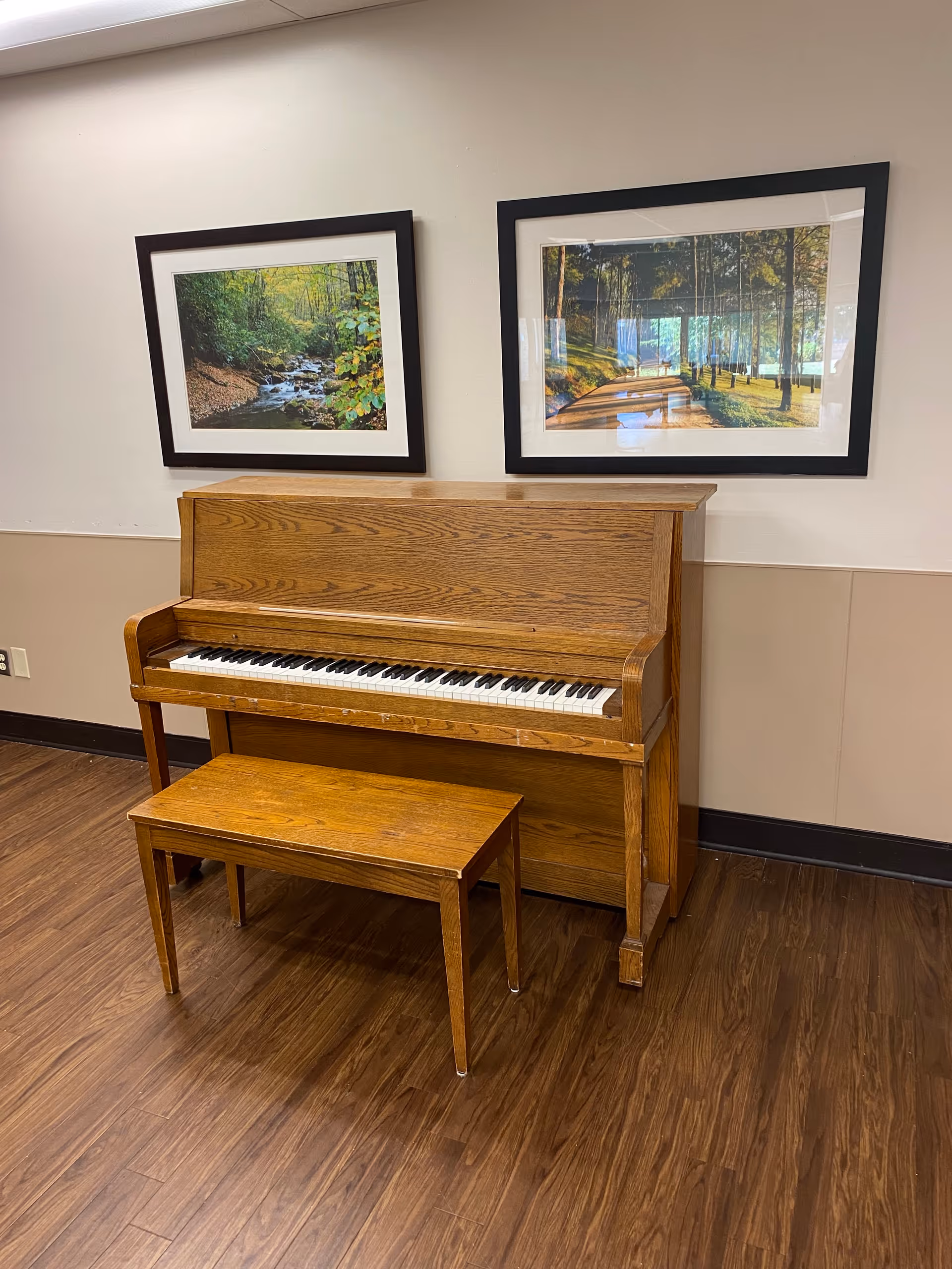 Wooden upright piano and bench against a beige wall with two framed landscape pictures above on a wood floor.