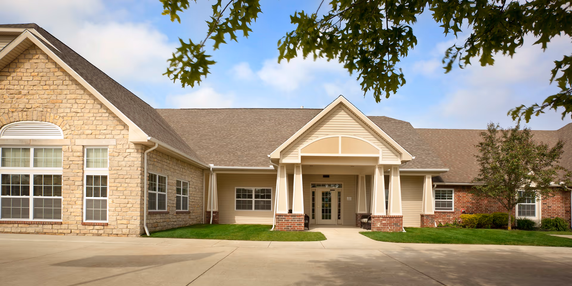 Front exterior view of a single-story senior living facility building with a covered entrance supported by columns, stone and brick facade, multiple windows, green lawn, and a tree branch with leaves in the foreground under a partly cloudy sky.