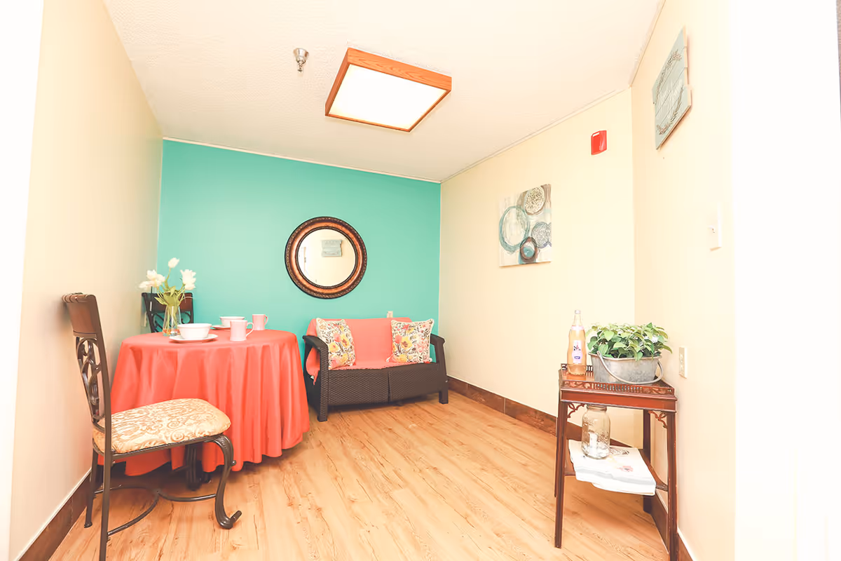 A small sitting area with a round table covered with a red tablecloth, two chairs, and a loveseat with floral cushions. The wall behind the loveseat is painted teal and has a round mirror. The adjacent wall is beige with a decorative painting and a small wooden side table holding a plant and a bottle.