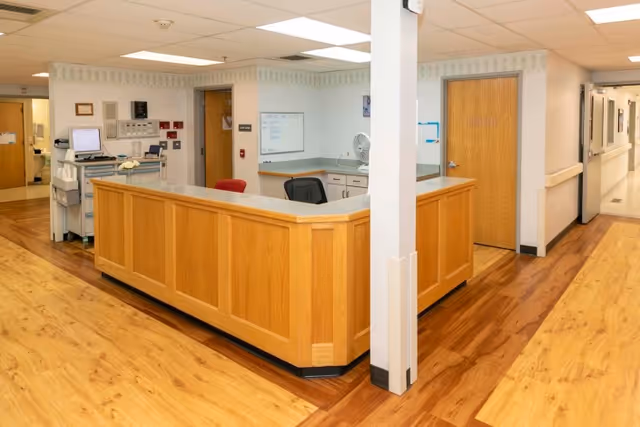Interior view of a nursing station in a rehabilitation and care center with a wooden counter, office chairs, a computer workstation, and doors leading to other rooms. The floor is wood-patterned, and the walls are light-colored with a subtle wallpaper border near the ceiling.