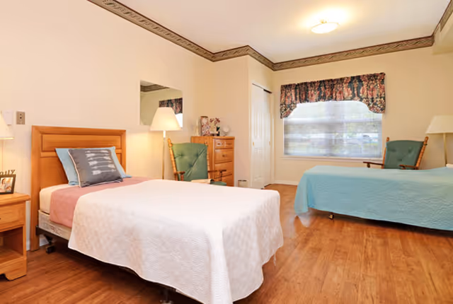 Bright shared bedroom with two single beds, wooden furniture, upholstered chairs, a window with a floral valance, and wood-look flooring.