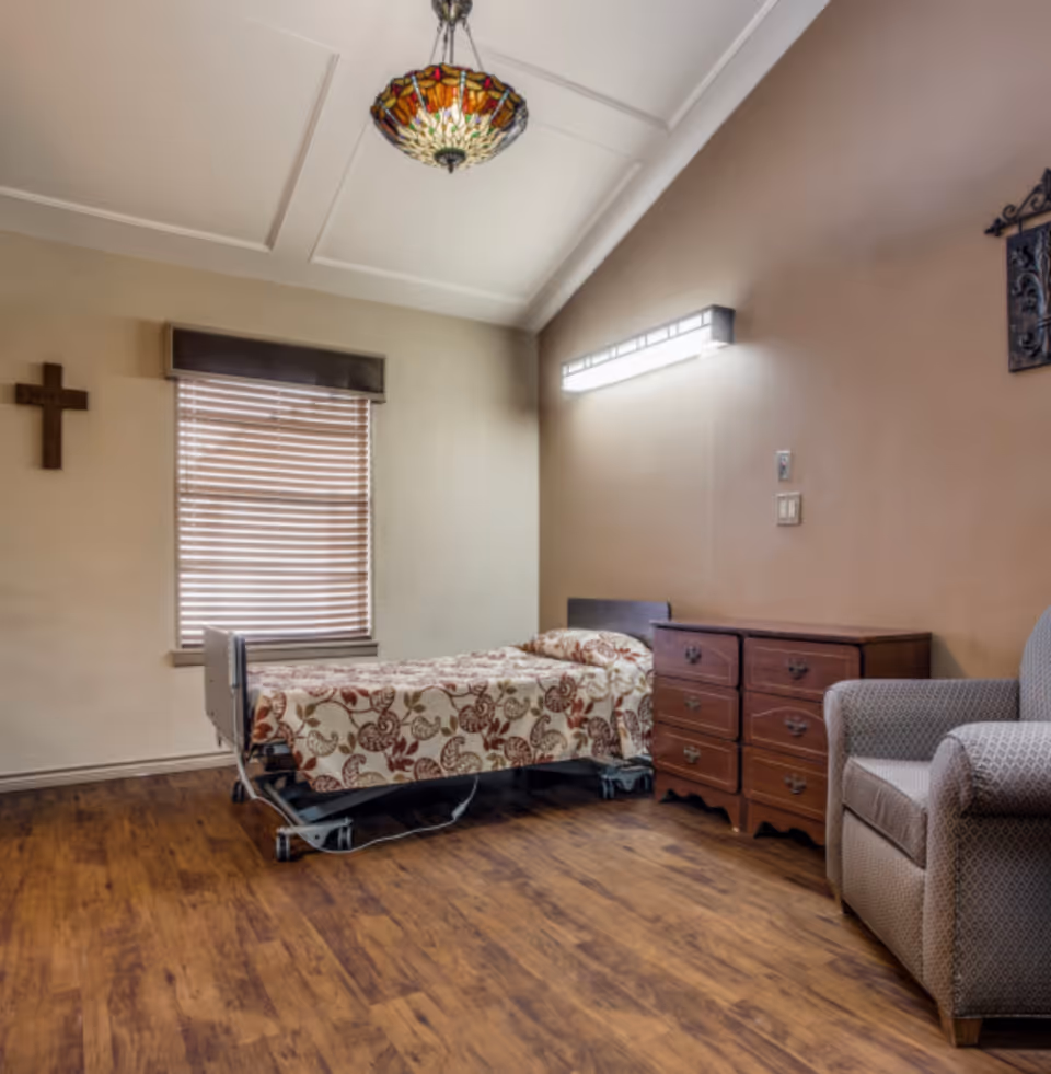 A simple bedroom in a care facility with a single bed covered in a floral patterned blanket, a wooden dresser with six drawers, a cushioned armchair, a window with closed blinds, a cross hanging on the wall, and a decorative ceiling light fixture.