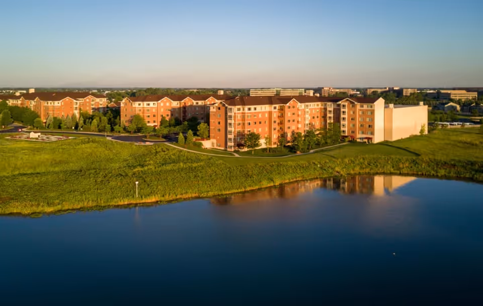 Aerial view of a large senior living facility named Sedgebrook, featuring multiple connected brick buildings with several floors, surrounded by green lawns and trees, with a body of water in the foreground reflecting the buildings.