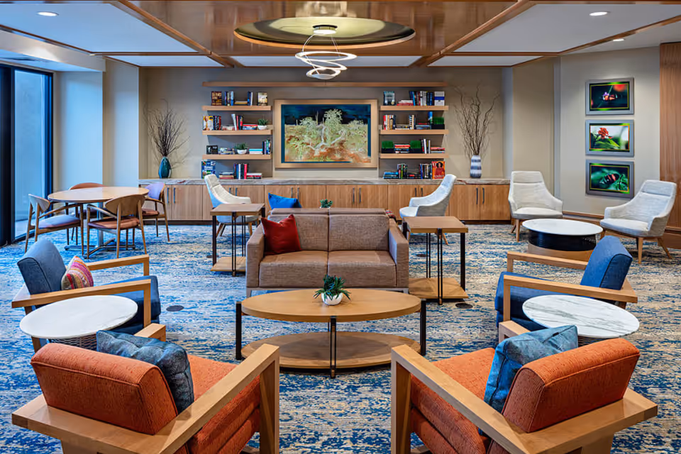 A spacious and modern living room area in a senior living facility with multiple seating options including armchairs with cushions, a sofa with pillows, and small round tables. The room features a blue patterned carpet, wooden furniture, built-in shelves with books and decorative items, and framed nature photographs on the wall. Large windows allow natural light to brighten the space.