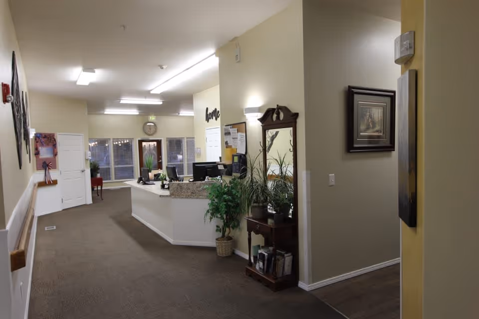 Interior view of a reception area in a senior living facility with a front desk, computer monitors, plants, a wall clock, and decorative wall art. The space has beige walls, carpeted floors, and fluorescent ceiling lights.