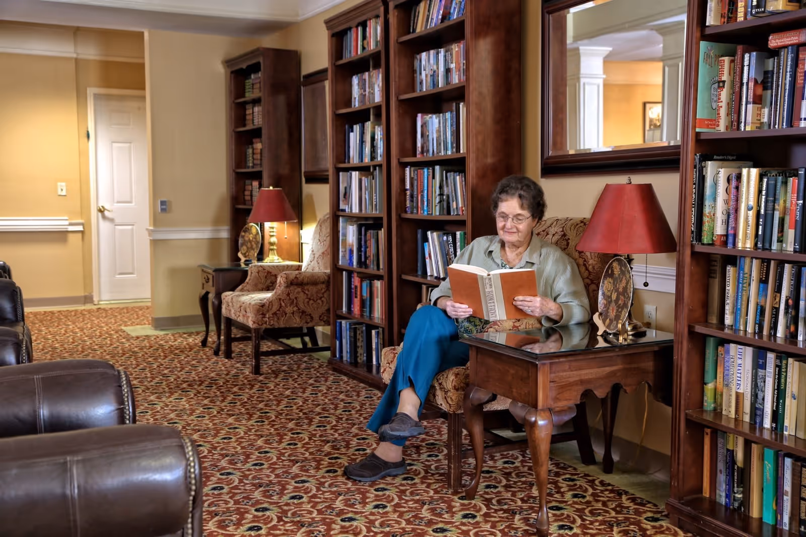 An elderly woman sits in a library-style common room reading a book beside bookshelves and a lamp.