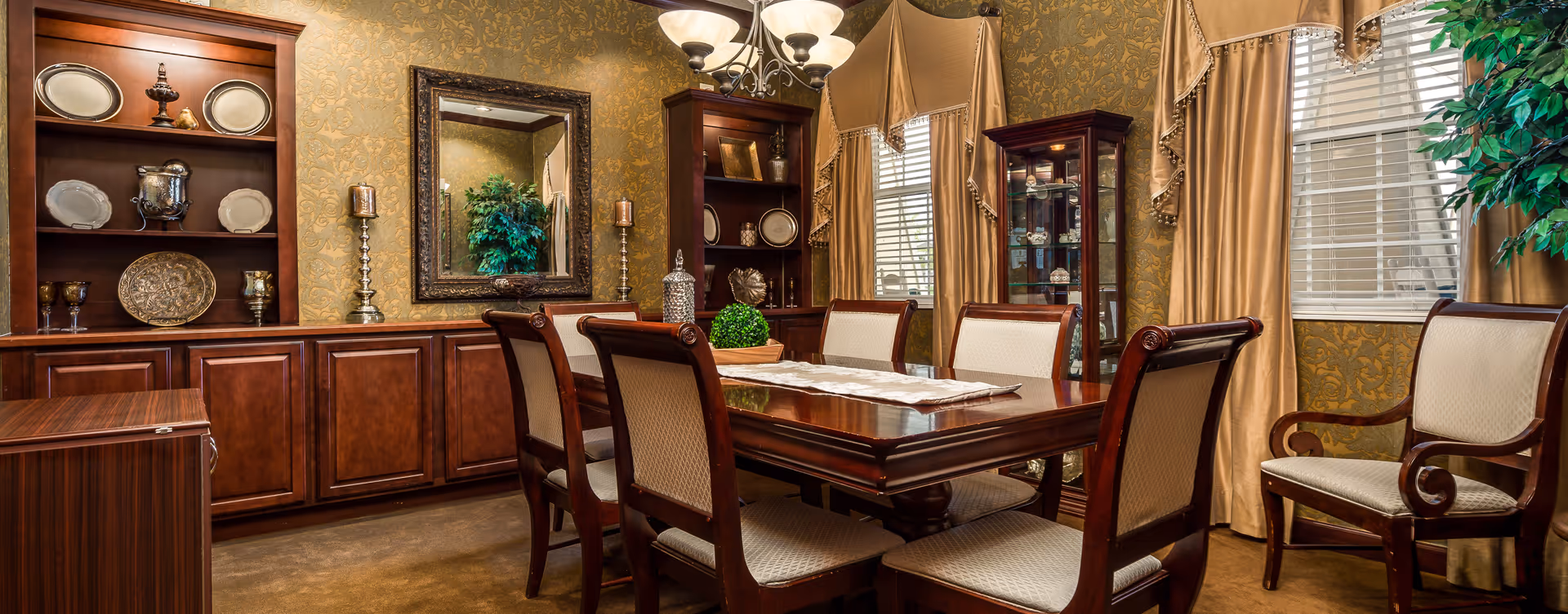 A formal dining room with a polished wooden dining table surrounded by six upholstered chairs. The walls are decorated with ornate gold-patterned wallpaper and two large windows with beige curtains. There are wooden cabinets and shelves displaying decorative plates, glassware, and ornamental items. A large mirror hangs on one wall, reflecting a green potted plant. A chandelier with multiple lights hangs above the table.