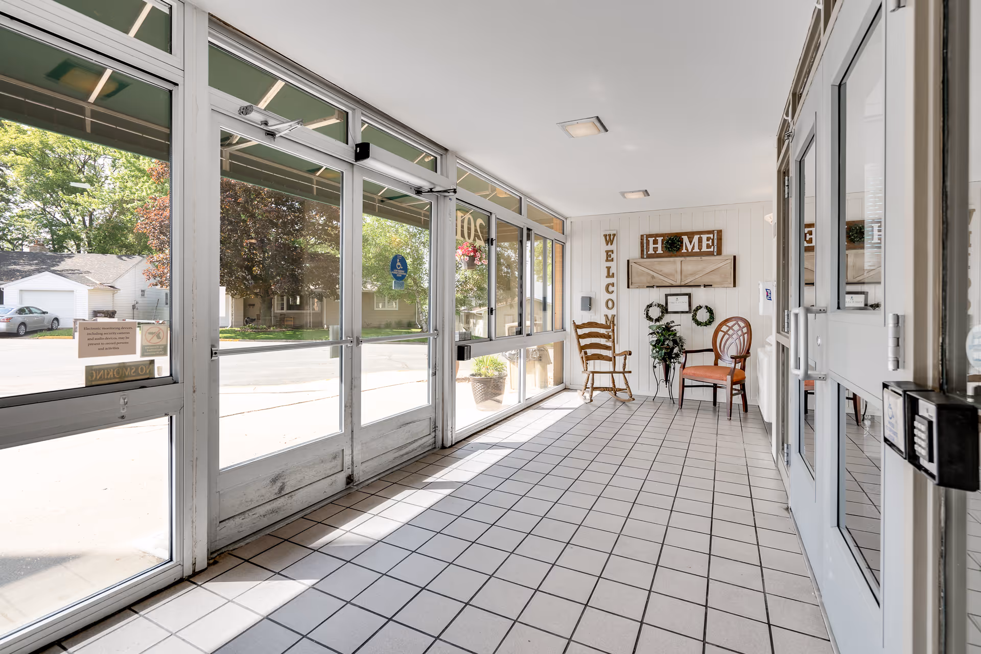 Bright entrance area with large glass doors and windows letting in natural light. The floor is tiled with light-colored square tiles. On the right side, there are two chairs, a small table with a plant, and wall decorations including a vertical 'WELCOME' sign and a 'HOME' sign with wreaths.