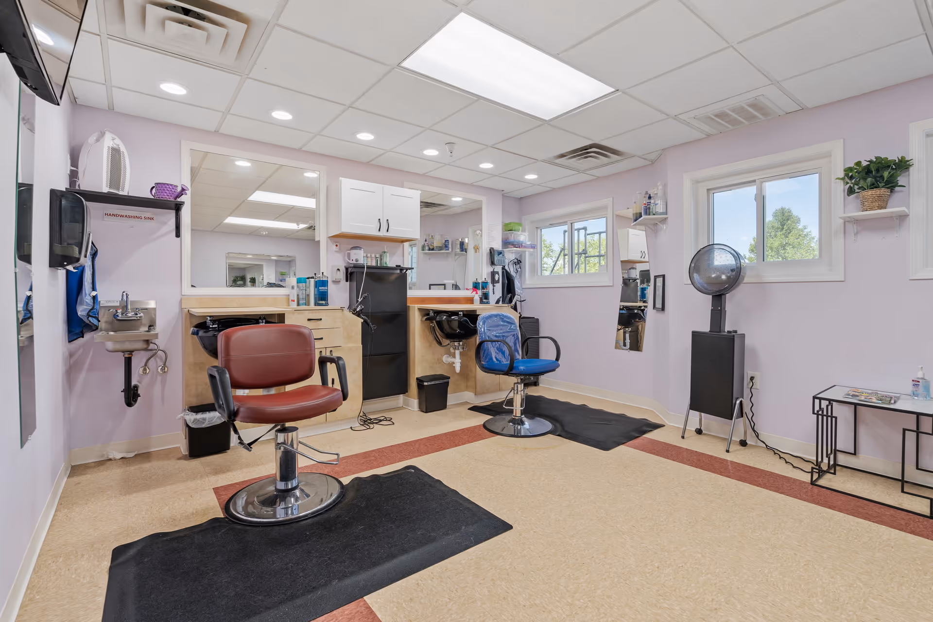Interior of a hair salon area in a senior living facility with two salon chairs, two hair washing sinks, large mirrors, a hair dryer, and shelves with hair care products. The walls are painted light purple and there are two windows letting in natural light.