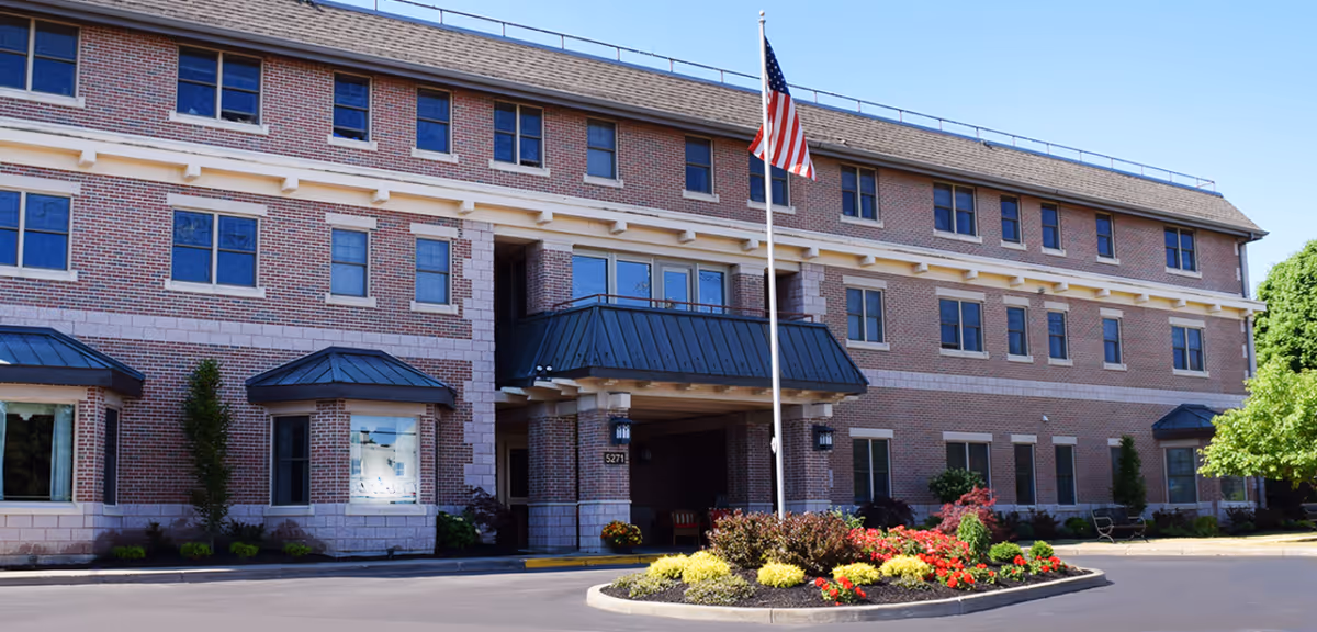 Front exterior view of a three-story brick senior living facility building with multiple windows, a covered entrance, an American flag on a flagpole, and a landscaped roundabout with colorful flowers and shrubs.