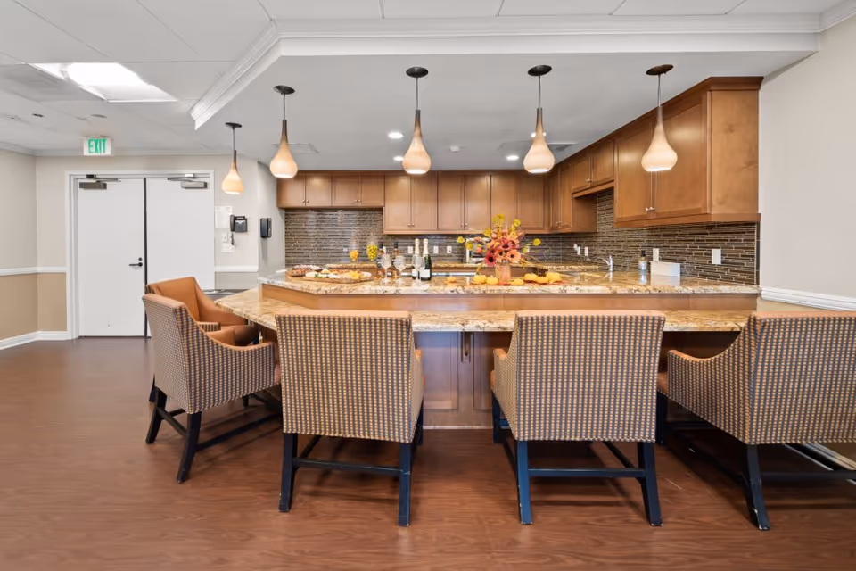 Spacious communal kitchen with a large granite island, pendant lights, wooden cabinets, and upholstered bar chairs.
