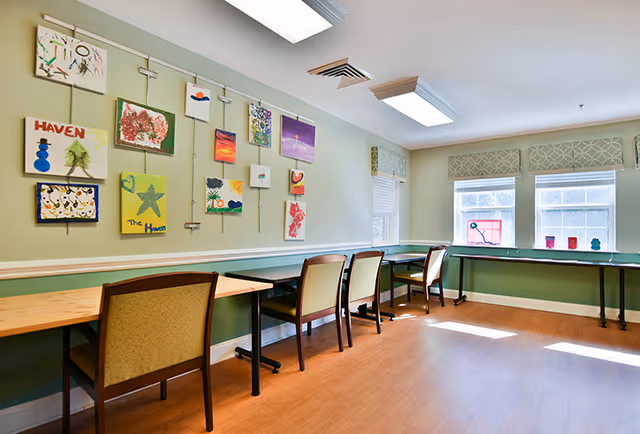 A well-lit room with light green walls featuring various colorful paintings and artwork displayed on the wall. There are several wooden tables with cushioned chairs arranged along the walls, and large windows with patterned valances letting in natural light. The floor is a light wood laminate.