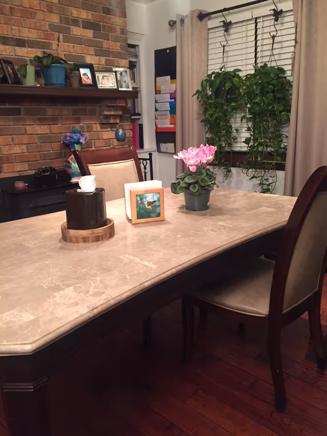 A dining room with a marble-top table and two wooden chairs with beige cushions. On the table are a tissue box, a small framed picture, and a potted pink flower. Behind the table is a window with beige curtains and two hanging green plants. To the left, there is a brick wall with a wooden mantel displaying framed photos and small decorative items.