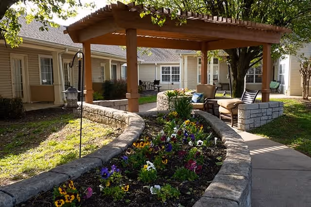 Outdoor garden area at Brookdale Gallatin featuring a raised stone flower bed with colorful flowers, a wooden pergola with seating underneath, and a paved walkway surrounded by trees and building windows.