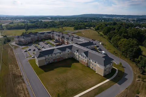 Aerial view of a large senior living facility named Harmony at Hershey, surrounded by green fields and trees, with multiple parking areas and a winding driveway.
