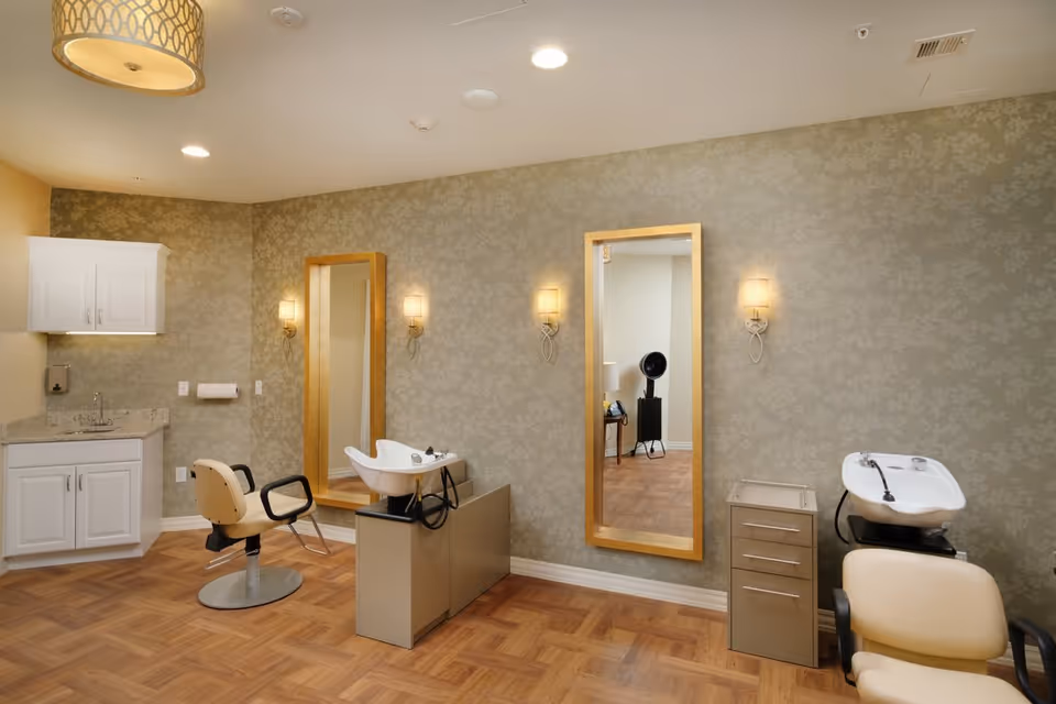 Well-lit salon room with two styling chairs, shampoo sinks, large mirrors, cabinets, and patterned wallpaper.