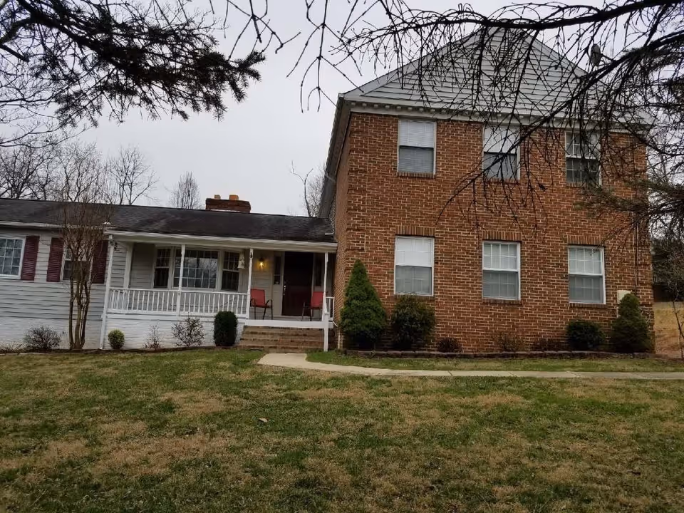 Front exterior view of a two-story assisted living facility building with a brick facade on the right side and white siding on the left side. There is a small porch with two red chairs and a lit porch light. The lawn in front is green with some patches of brown, and there are trees and shrubs around the building under an overcast sky.