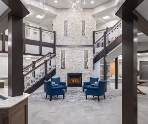 A spacious and modern common area with a double staircase on either side leading to an upper level. The focal point is a tall stone fireplace with a fire burning, flanked by two vertical decorative panels. In front of the fireplace are four blue upholstered chairs arranged around a small table on a patterned rug. The area is well-lit with recessed lighting and has a neutral color palette with dark wood accents.