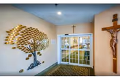 Interior view of a hallway entrance at St. John Neumann Center for Rehabilitation & Healthcare featuring a decorative metal tree wall art on the left wall, a crucifix mounted on the right wall, and glass double doors leading to another area.