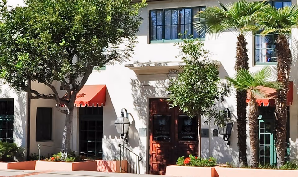Exterior view of Villa Santa Barbara featuring a white building facade with dark wooden double doors, red awnings over windows, and lush greenery including trees and palm plants in front.
