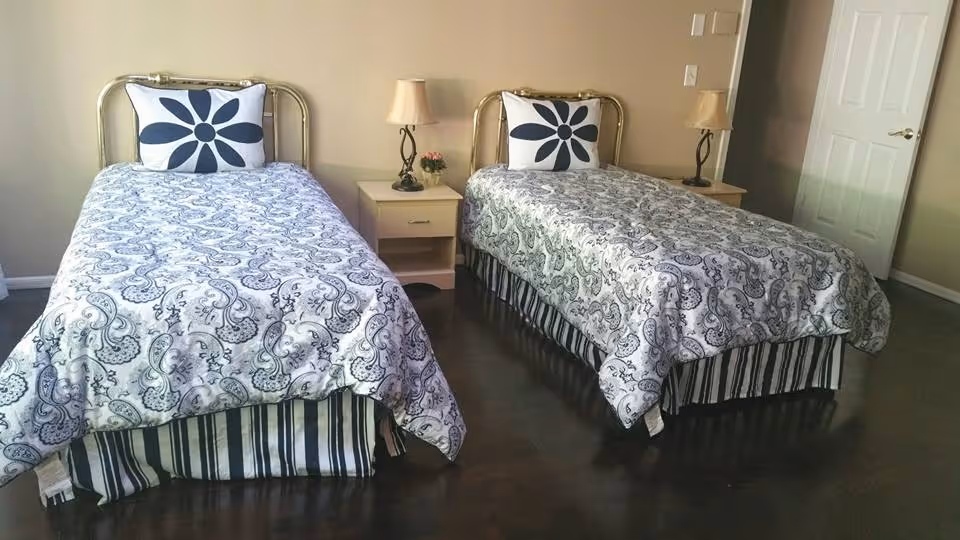 A bedroom with two twin beds, each with a white and black patterned bedspread and a pillow featuring a large black flower design. Between the beds is a small nightstand with a lamp and a small flower pot. The room has wooden flooring and beige walls, with a white door visible on the right side.