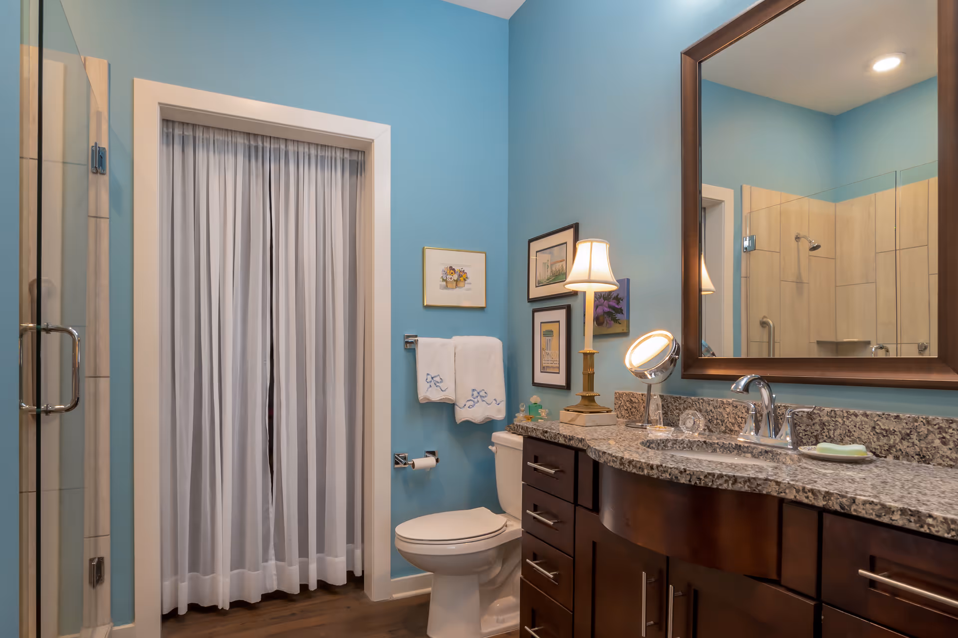 A bathroom with light blue walls featuring a granite countertop with a sink, a large mirror, a small lamp, and a round makeup mirror. There is a toilet with two white towels hanging above it, framed artwork on the wall, a glass-enclosed shower, and a doorway with sheer white curtains.