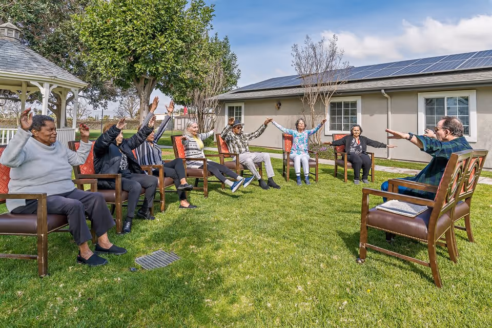 A group of elderly people sitting in a circle on chairs outdoors on a grassy lawn, participating in a seated exercise or stretching activity with their arms raised. There is a gazebo and trees in the background, along with a building with solar panels on the roof.