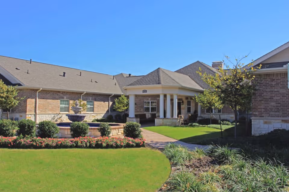 Exterior view of a brick senior living facility entrance with a central tiered fountain, covered portico with columns, and manicured lawns.