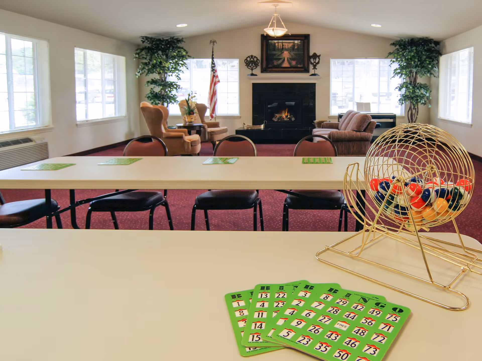A bright room with large windows and red carpet featuring tables and chairs arranged for a bingo game. On the table in the foreground are green bingo cards and a gold bingo cage filled with colorful bingo balls. In the background, there are comfortable armchairs and a sofa near a fireplace with a painting above it, flanked by two large potted plants and an American flag.