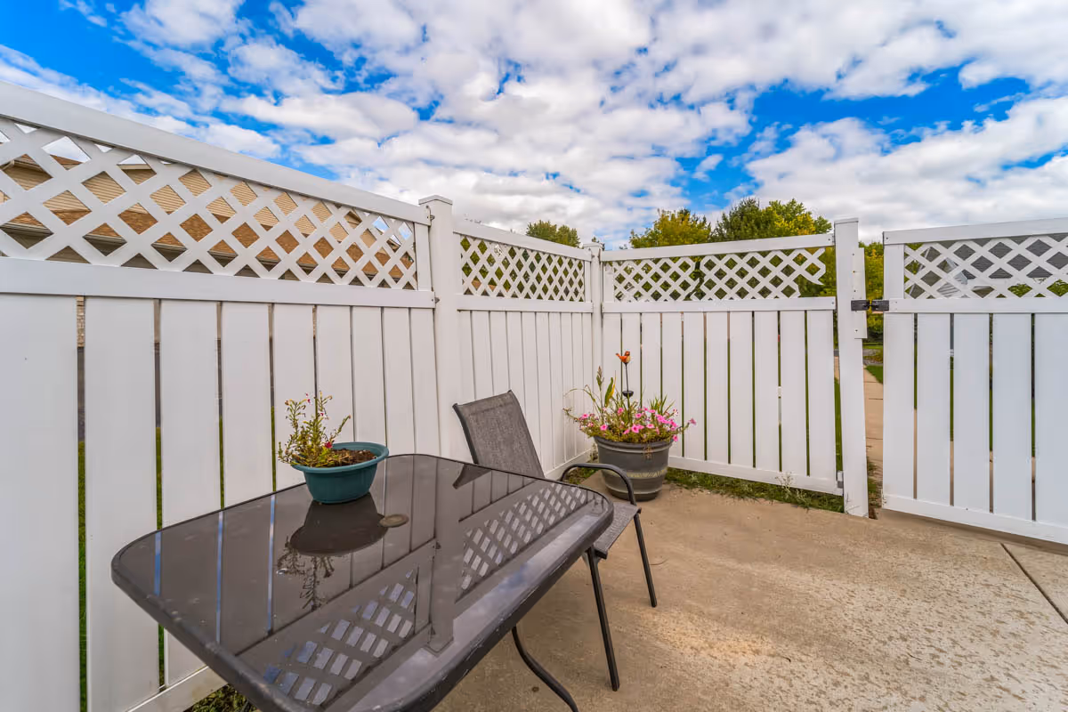 A small outdoor patio area enclosed by a white lattice fence with a gate. The patio has a glass-top table with a potted plant on it and a single chair. Another larger pot with flowers is placed near the corner of the fence. The sky is partly cloudy with patches of blue visible.