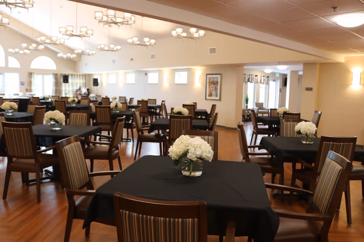 A spacious dining room with multiple square tables covered in black tablecloths, each adorned with a vase of white flowers. The room features wooden chairs with striped upholstery, warm wooden flooring, modern chandeliers hanging from the ceiling, and large windows allowing natural light to fill the space.