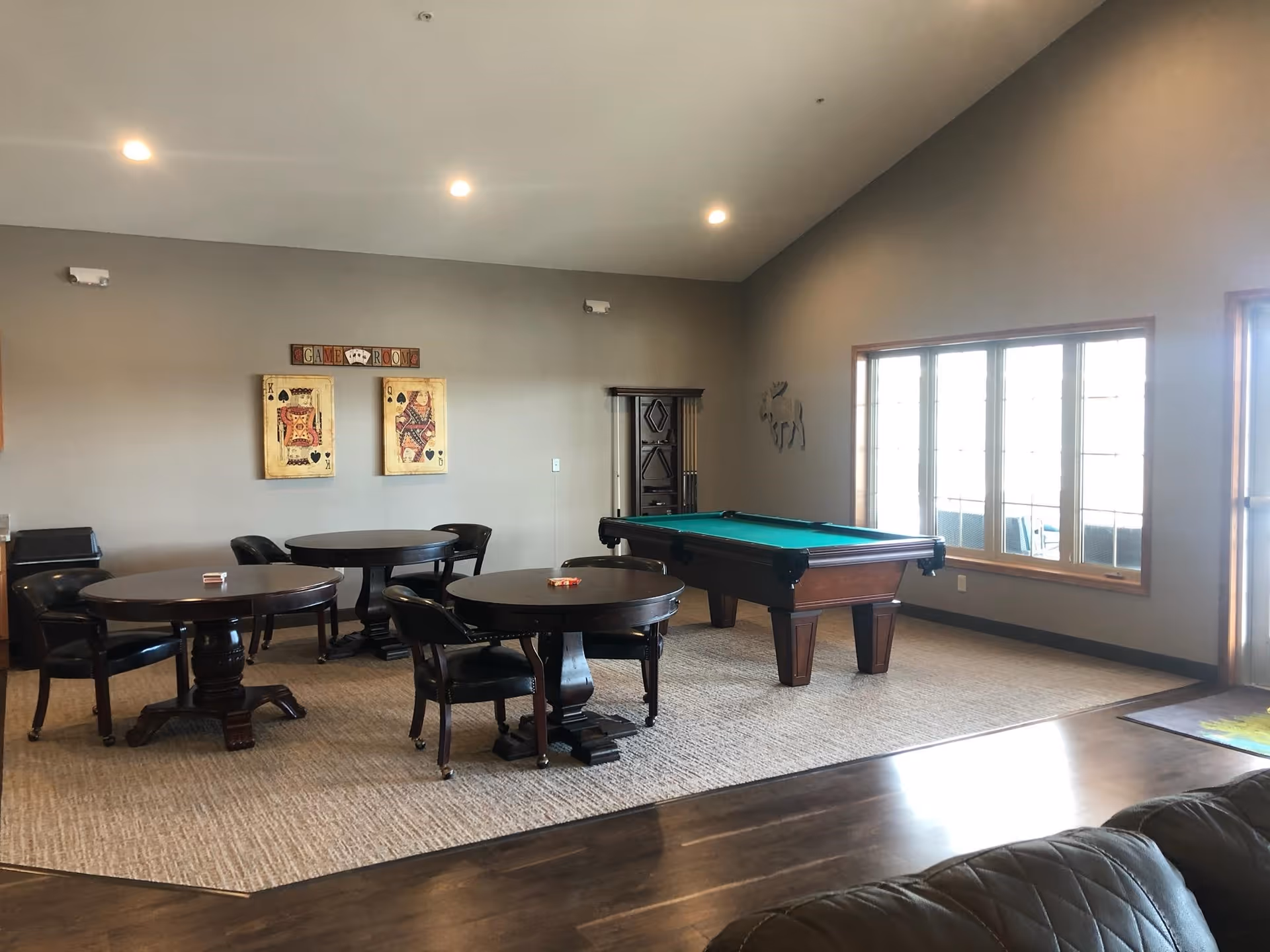 A spacious game room with three round tables and chairs, a pool table, and large windows letting in natural light. The walls are decorated with playing card artwork and a sign that says 'Game Room'. The floor is a combination of carpet and wood.