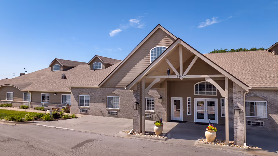 Front exterior of a single-story assisted living building with a covered gabled entrance, double glass doors, and potted plants.