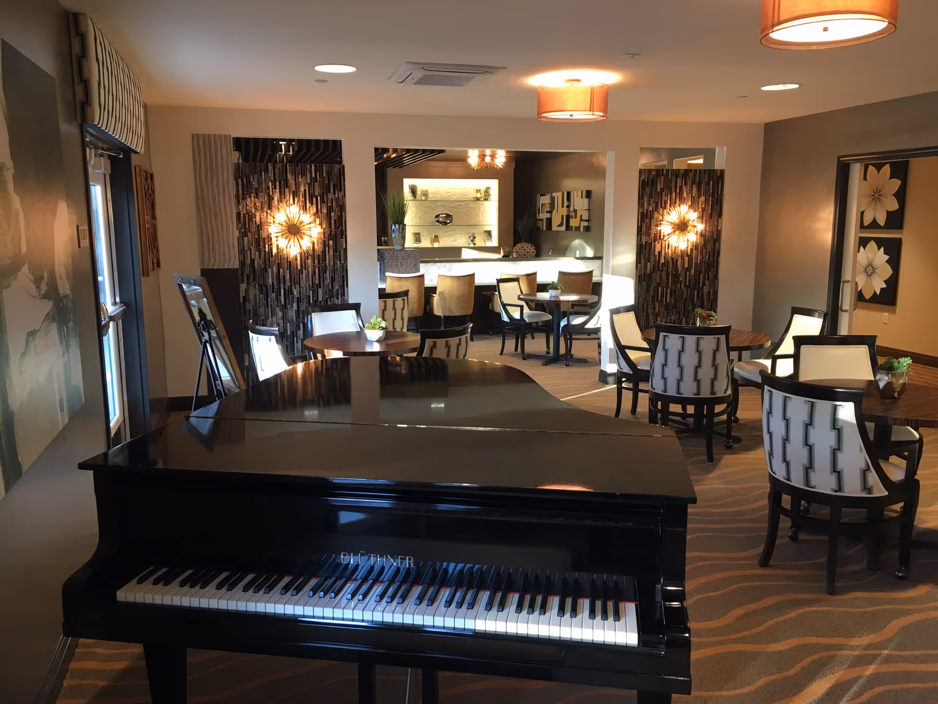 Interior view of a common area in a senior living facility featuring a black grand piano in the foreground, several round tables with cushioned chairs, decorative wall art, and warm lighting fixtures.