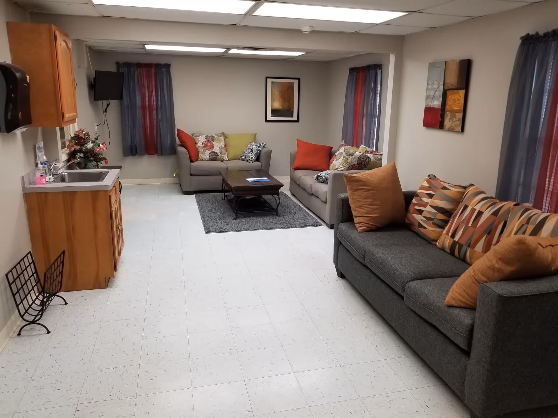 A communal living room with gray sofas and colorful throw pillows arranged around a coffee table, a small sink and wooden cabinet on the left, and framed artwork on the walls.